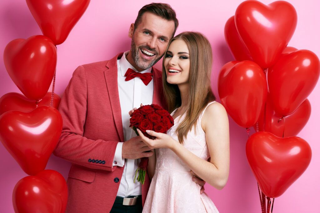 Casal feliz se abraçando e a mulher procura namorado com buquê de rosas vermelhas e um ambiente decorado com balões em forma de coração