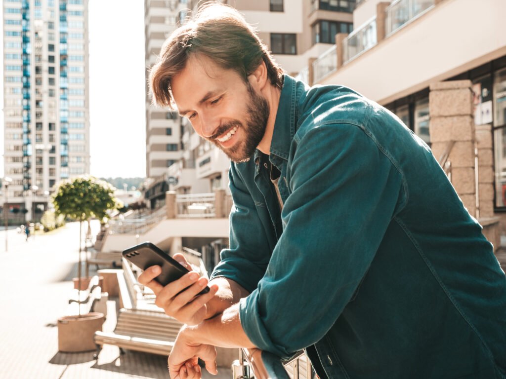 Homem branco, de barba usando camisa social jeans e olhando sorridente seu celular